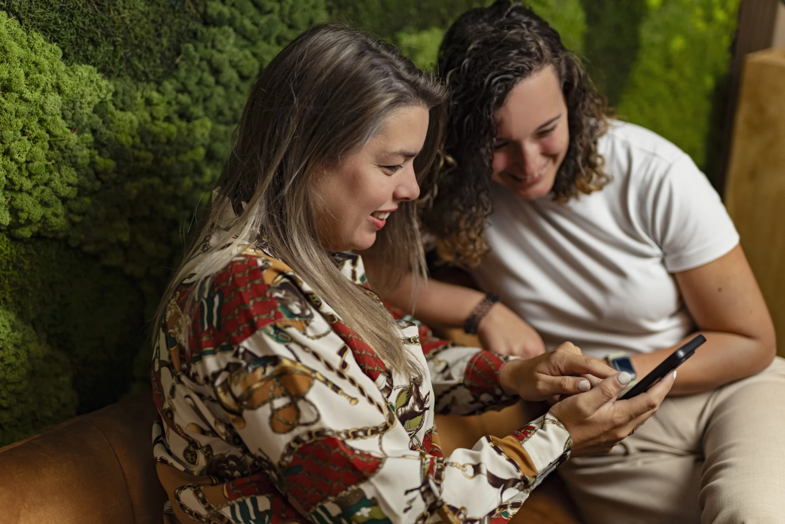 Twee vrouwen op een bankje, met een groene achtergrond met een telefoon met een blij gezicht.